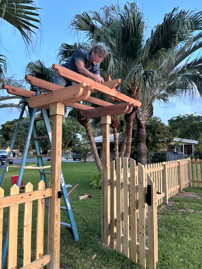 Worker atop trellis structure with picket fence in residential yard
